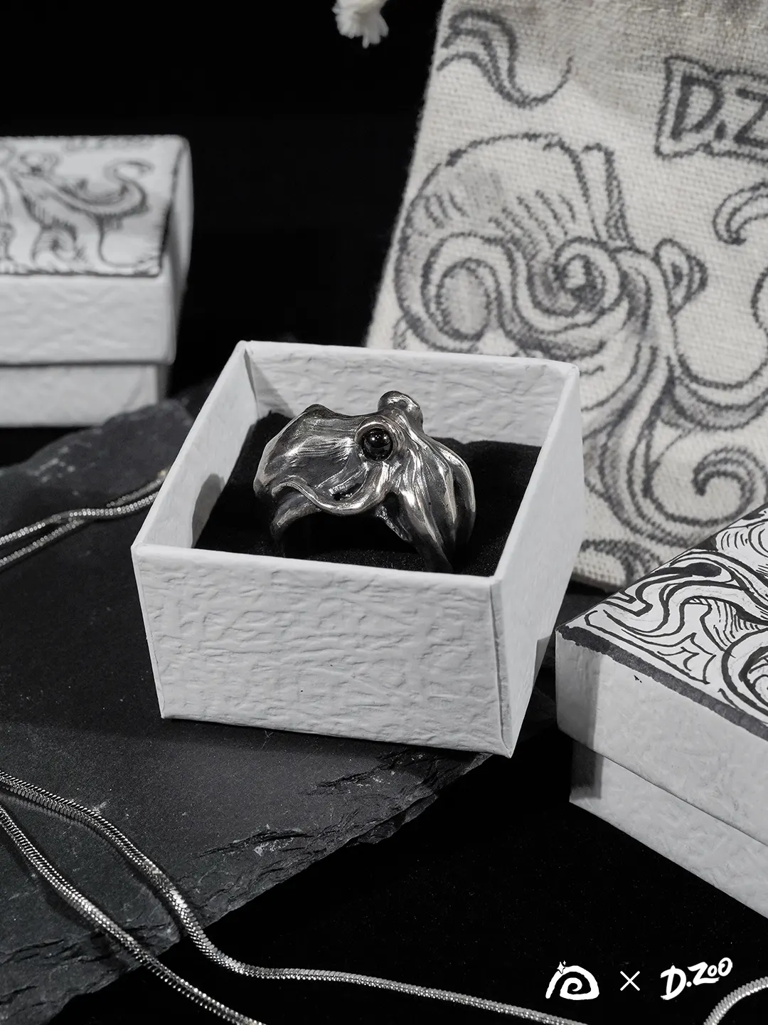 A silver octopus ring displayed inside a hand-drawn octopus jewelry box with a fabric pouch in the background.
