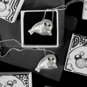 Two sterling silver Ploop seal necklaces displayed with hand-drawn jewelry boxes, one resting on a black stone slab and one nestled inside a box.