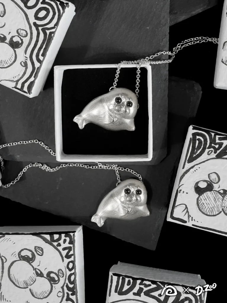 Two sterling silver Ploop seal necklaces displayed with hand-drawn jewelry boxes, one resting on a black stone slab and one nestled inside a box.