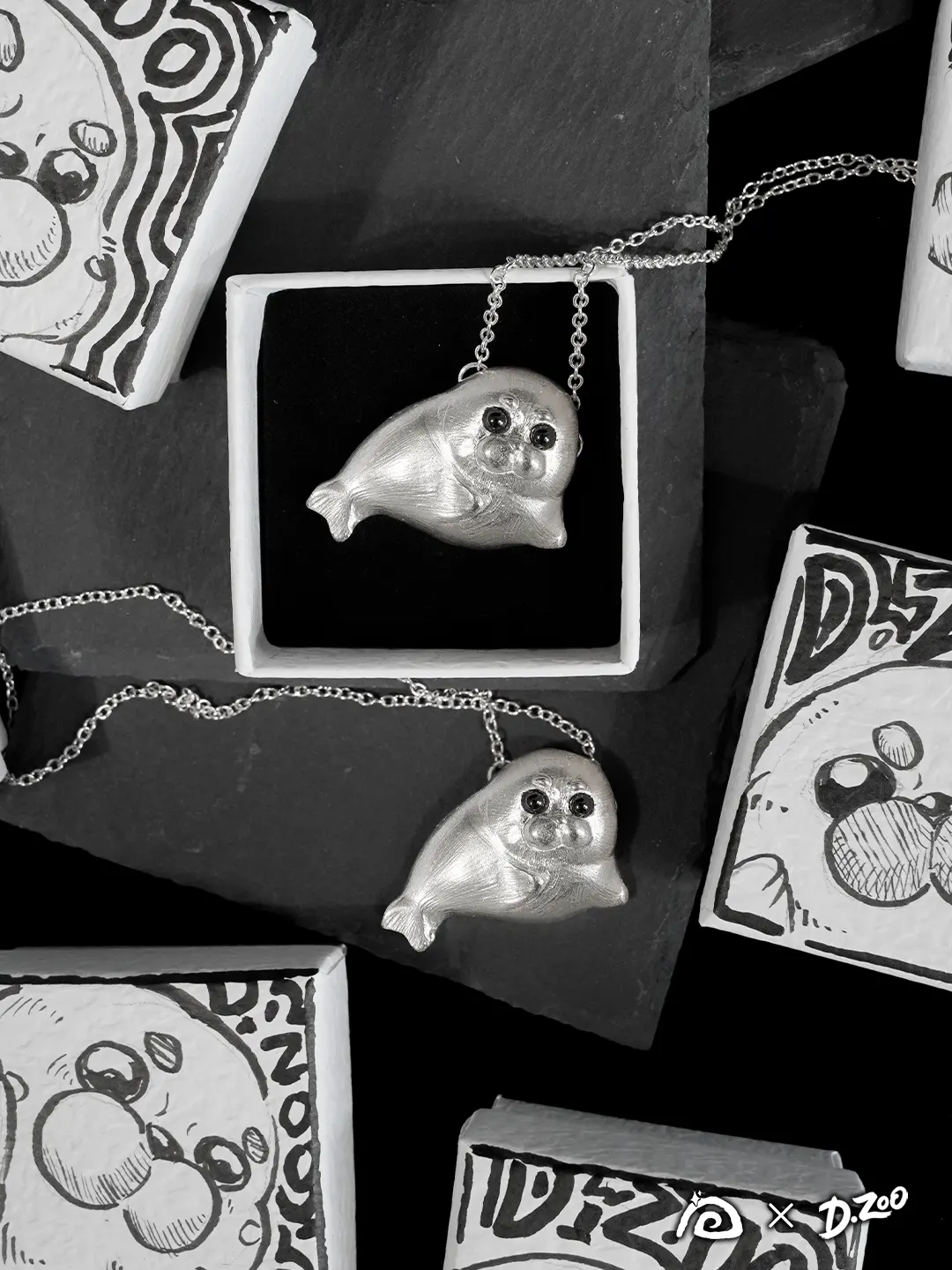 Two sterling silver Ploop seal necklaces displayed with hand-drawn jewelry boxes, one resting on a black stone slab and one nestled inside a box.