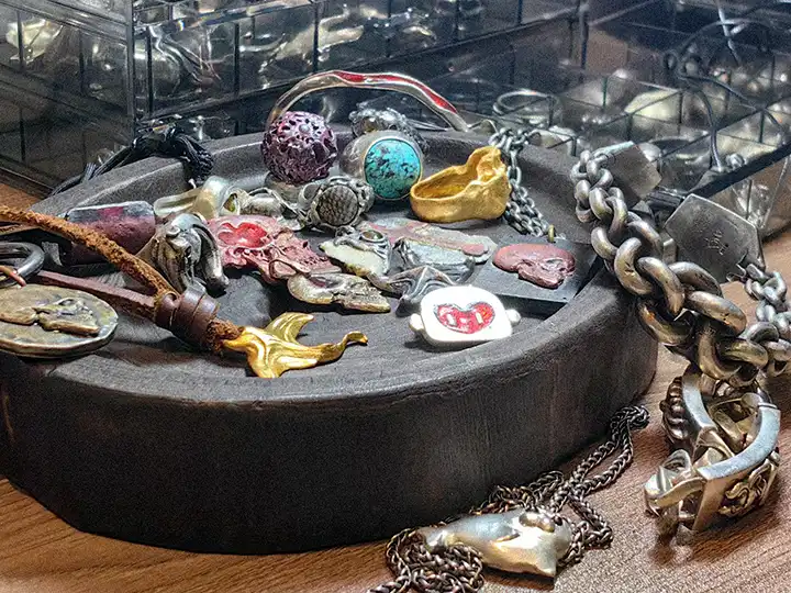 A collection of handcrafted artistic jewelry pieces displayed on a round wooden surface, with additional small creations arranged inside a clear acrylic box in the background, representing Rainbow Tears' Petite Rarities series.