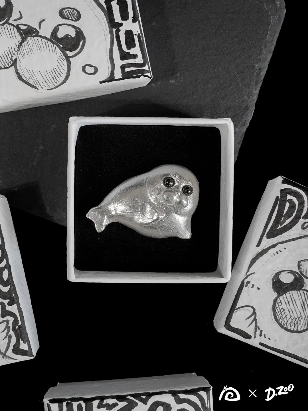 A rounded sterling silver seal brooch resting in a jewelry box, surrounded by four hand-drawn illustrated gift boxes.