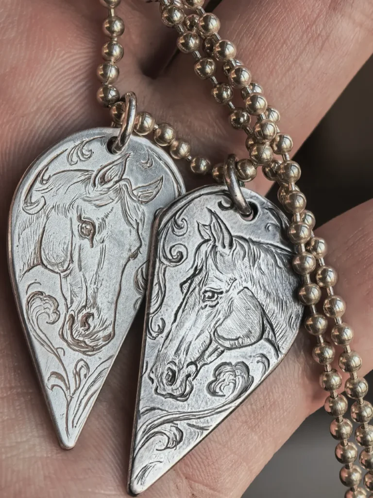 A pair of 990 fine silver half-heart pendants being held in hands, featuring custom hand-engraved horse motifs that form a complete heart when joined together.