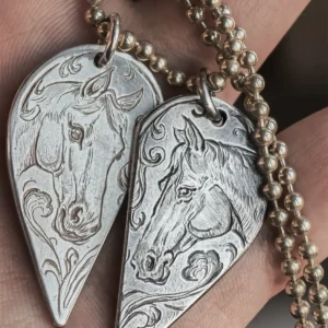 A pair of 990 fine silver half-heart pendants being held in hands, featuring custom hand-engraved horse motifs that form a complete heart when joined together.