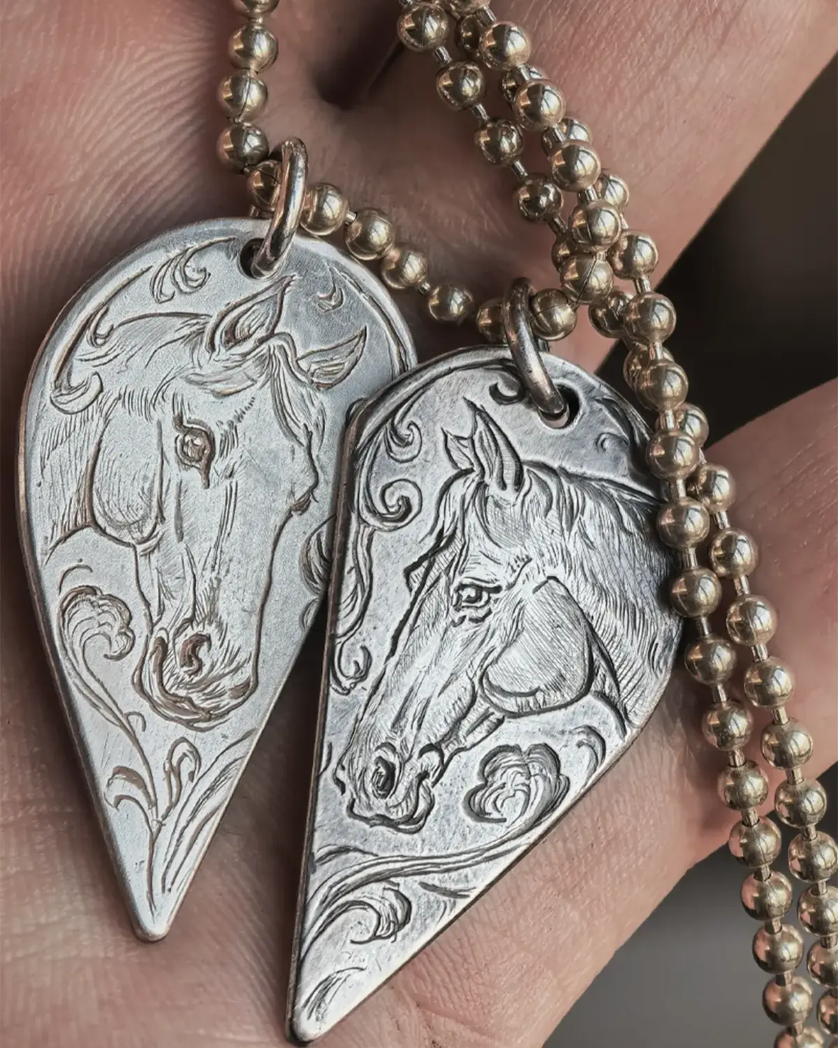 A pair of 990 fine silver half-heart pendants being held in hands, featuring custom hand-engraved horse motifs that form a complete heart when joined together.