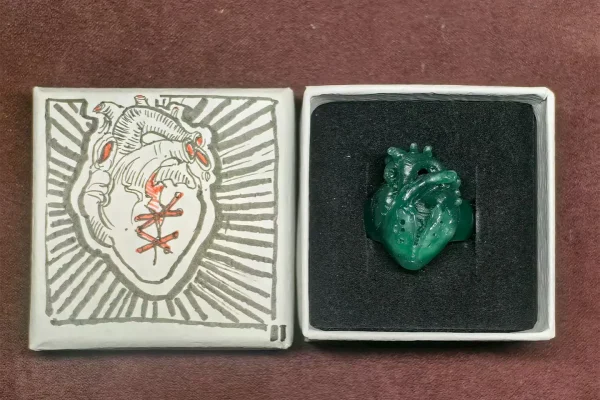 A hand-carved wax model for a split-heart ring resting in an open gift box, featuring a designer's custom-painted heart motif on the inner lid. This captures the artisanal jewelry-making process from concept to prototype.